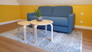 Una habitación con un sofá azul, dos mesitas redondas, un tapete floral y una planta en maceta.
