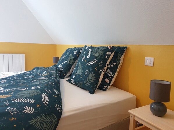 A bed with a dark green floral bedspread and matching pillows, a white mattress, and a small bedside table with a lamp.