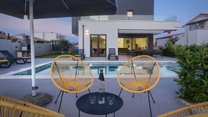 Een modern buitenzwembad met twee rieten stoelen en een tafel, een overdekte parasol en uitzicht op een modern huis met glazen deuren.