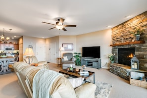 A living room with a fireplace, a television, a ceiling fan, and a kitchen area in the background.