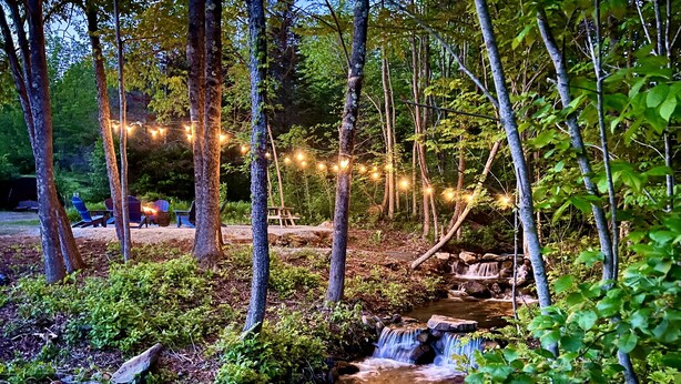 Zona boscosa con una pequeña cascada, luces colgantes y mobiliario exterior.