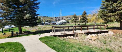 A wooden bridge over a concrete path in a park with boats in the background.