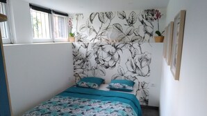 Un dormitorio pequeño y ordenado con un papel tapiz de motivos florales, una cama con ropa de cama color turquesa y una ventana con persianas.