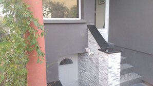 Una escalera con barandilla negra que conduce a una puerta, una ventana pequeña y una planta con flores.