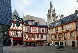 Ein Kopfsteinpflasterplatz mit traditionellen Fachwerkhäusern, einem Kirchturm und einer Bank.