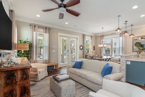 A spacious living room with a sofa, ottoman, and coffee table. There is a dining area with a table and chairs. The kitchen has a blue countertop and pendant lights.