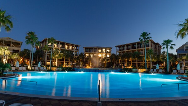 A large outdoor swimming pool surrounded by palm trees and a multi-story hotel building with balconies.