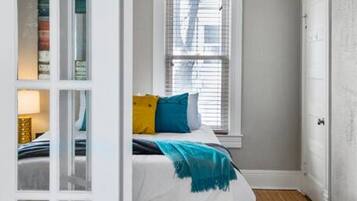 Une chambre avec un lit, une fenêtre avec des stores, un plancher en bois et une porte blanche.