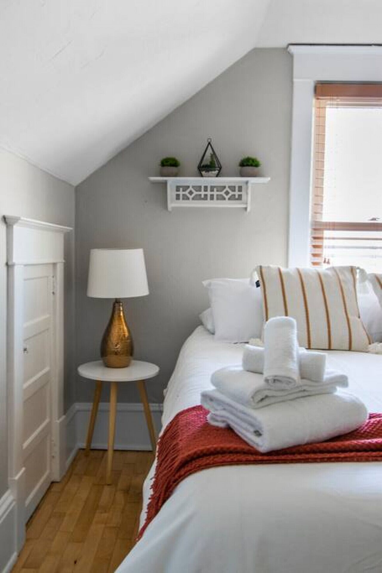 Un dormitorio con cama, mesita de noche con lámpara, estante con objetos decorativos y ventana con persianas.