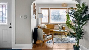 A living room with a sofa, armchair, and coffee table, a large potted plant, and a view through an arched doorway.
