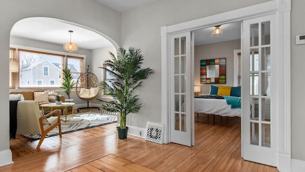 A spacious living room with hardwood floors, a large potted plant, a hanging chair, and a dining area with a patterned rug.