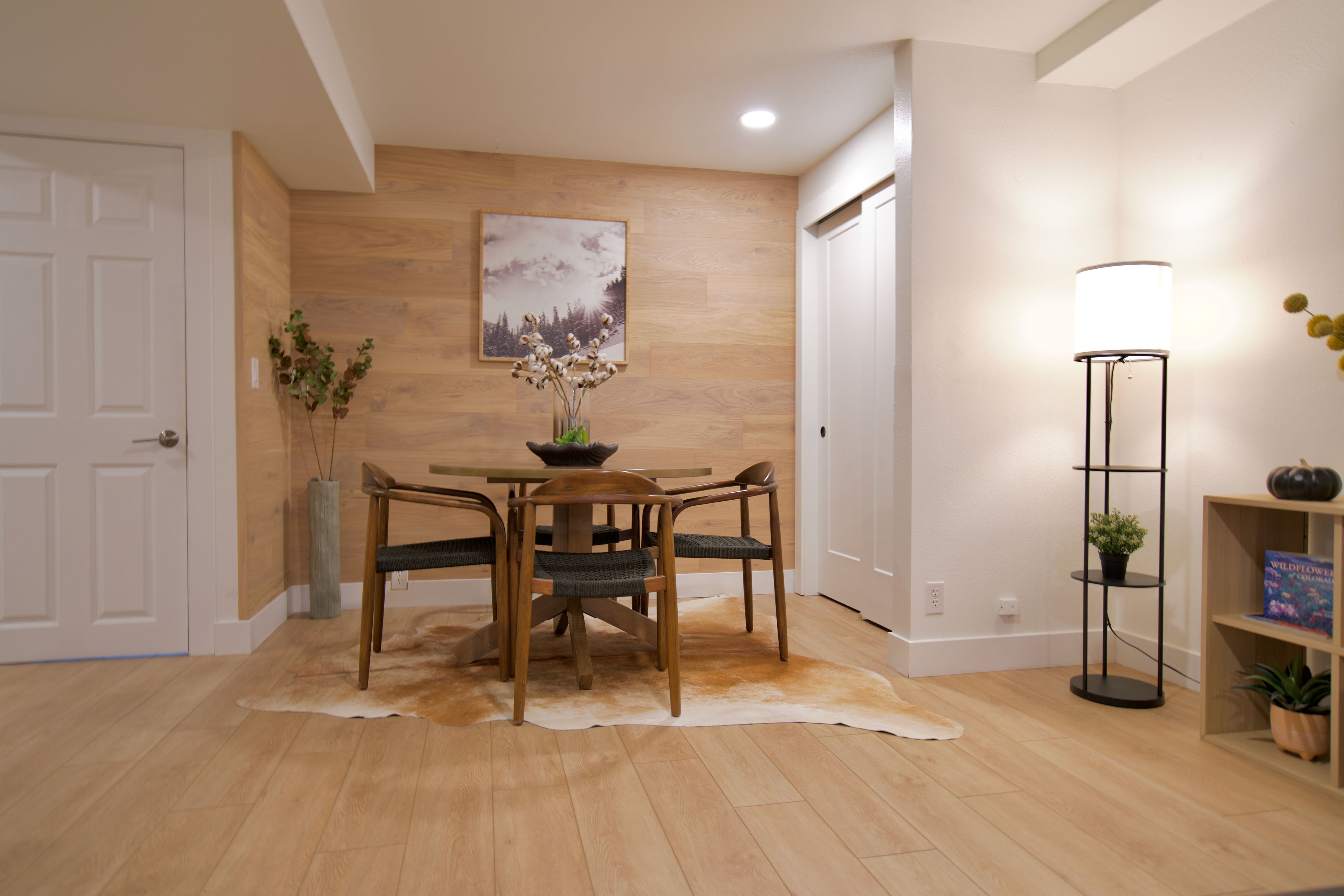 A dining area with a wooden table and chairs, a vase with flowers, a painting on the wall, a lamp, and a bookshelf with books.