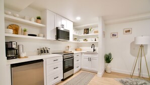 Una cocina moderna con armarios blancos, horno empotrado, microondas y lavavajillas. Hay estantes con objetos decorativos y una planta pequeña.