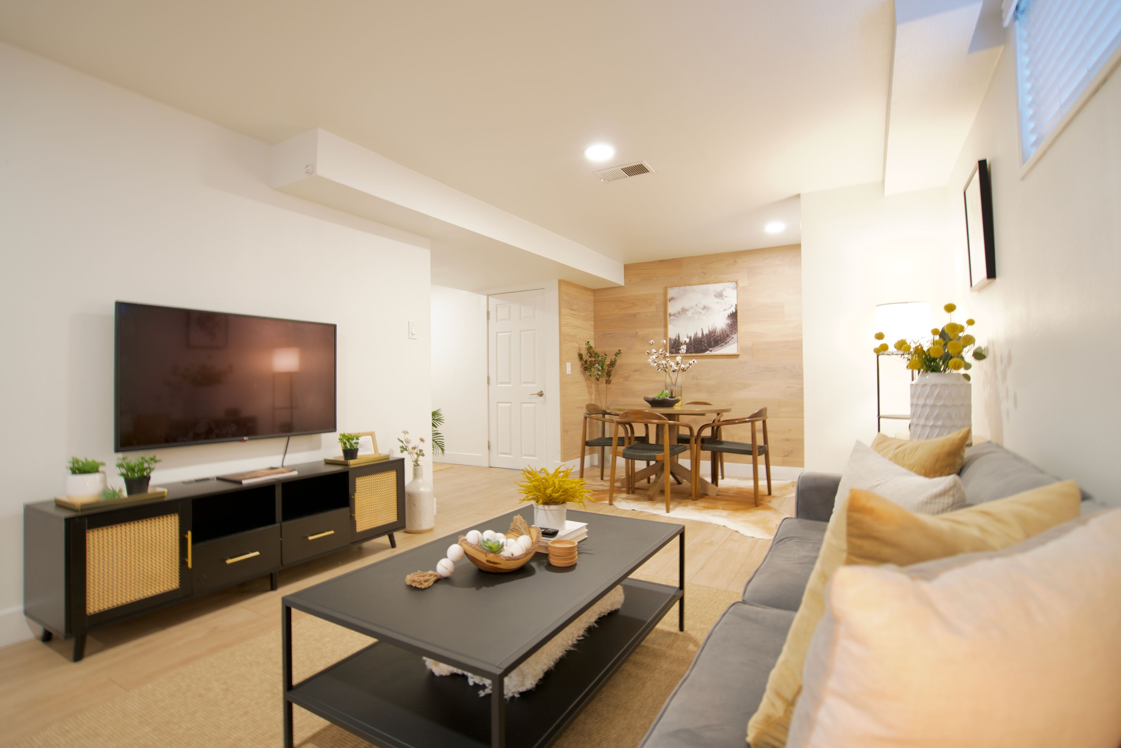 A modern living room with a flat-screen TV, a coffee table, a sofa, and a dining area with a wooden table and chairs.