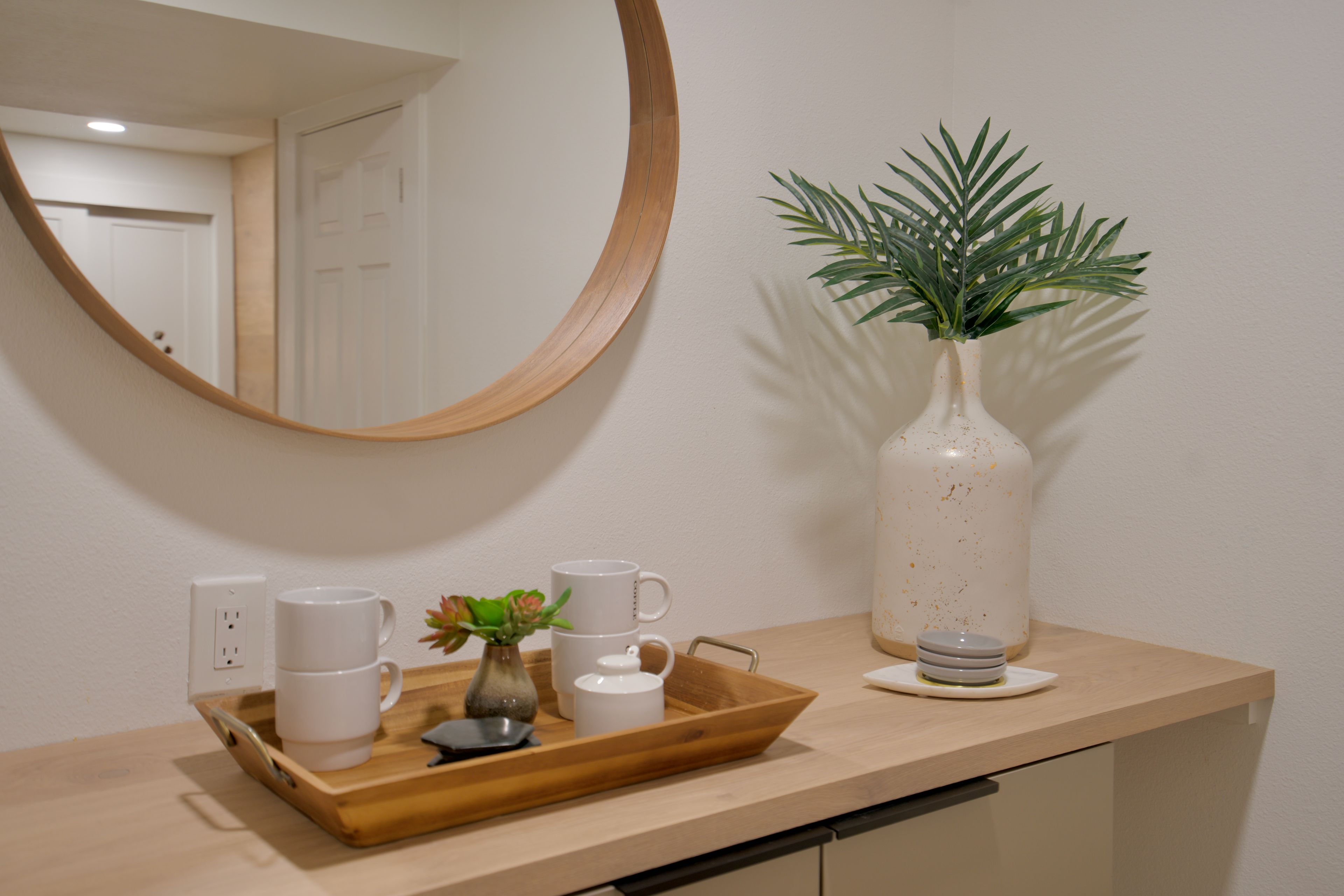 A kitchen counter with a wooden tray holding white mugs, a small vase with a plant, and a white pitcher.
