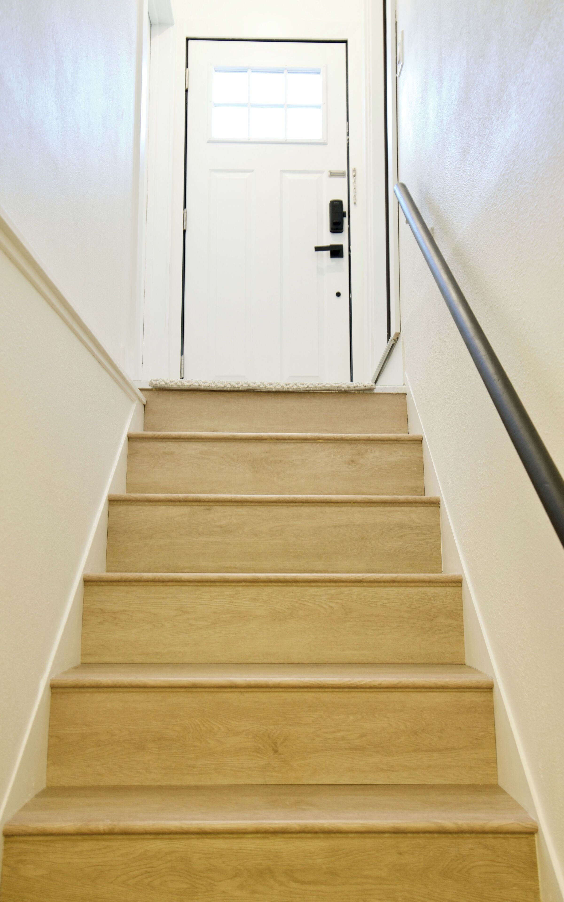 Una escalera que lleva a una puerta blanca con ventana, escalones de madera y barandilla de metal.