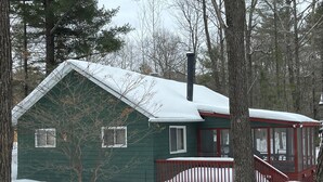 Une cabane verte avec un porche rouge et une cheminée, entourée d’arbres et recouverte de neige.