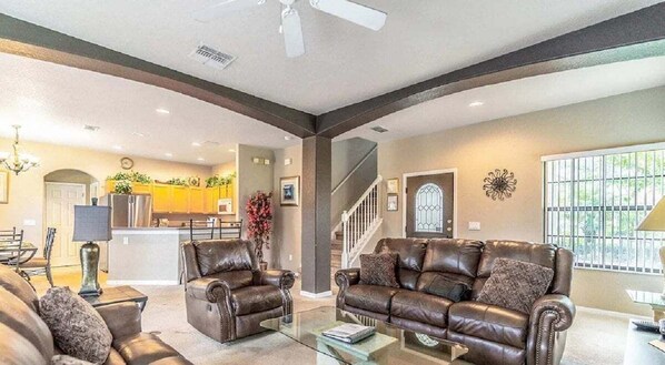 A spacious living room with leather sofas, a glass coffee table, and a kitchen in the background.