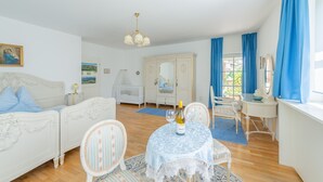 Ein Zimmer mit einem weißen Bett, einem Esstisch mit Stühlen, einem Spiegel und einem Fenster mit blauen Vorhängen.