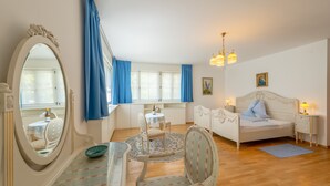 Ein Schlafzimmer mit einem Bett, Nachttischen, einem Schminktisch, einem Spiegel, einem Fenster mit Vorhängen sowie einem Essbereich mit Tisch und Stühlen.