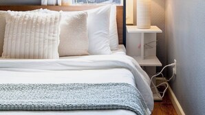 Un dormitorio con cama, ventana con cortinas, mesita de noche con lámpara y estantería con libros.