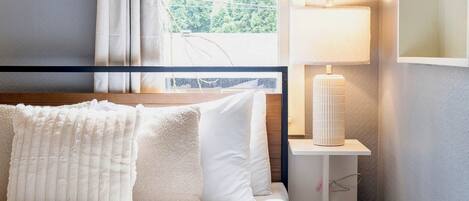 Un dormitorio con cama, ventana con cortinas, mesita de noche con lámpara y estantería con libros.