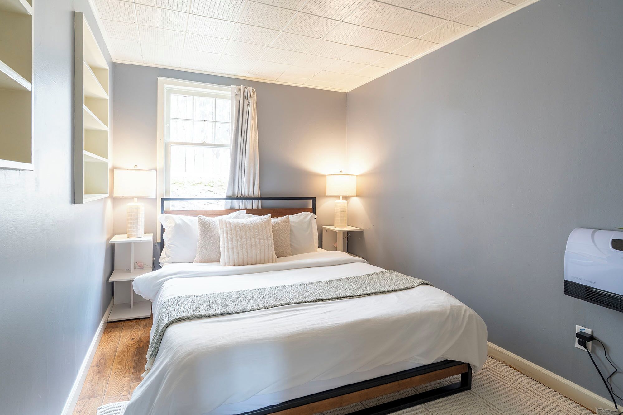 A neatly arranged bedroom with a bed, bedside lamps, a window with curtains, and a wall-mounted heater.