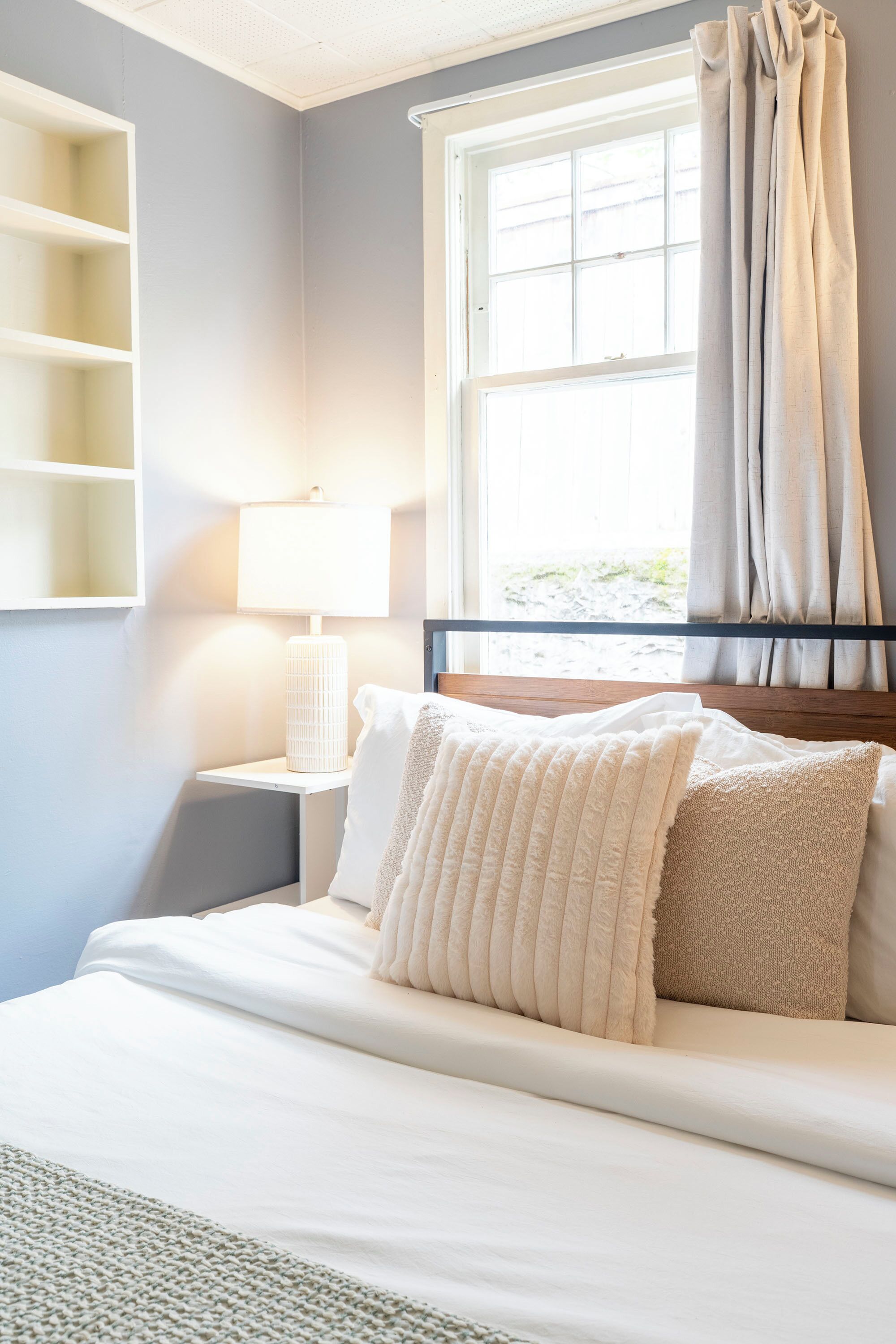 A neatly made bed with white linens and two pillows, a bedside lamp, and a window with curtains.