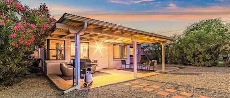 A covered outdoor patio with seating, a grill, and a gravel pathway leading to a house.