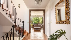 A modern interior with a herringbone wood floor, a black metal staircase, a chandelier, a large mirror, and a view into a living area with a sofa and plants. - Highgate - Luxury 4-Bedroom Spacious House with Indoor Pool (London)