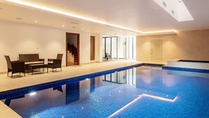 A modern indoor swimming pool with a clear glass ceiling, a seating area with a sofa and chairs, and a visible exercise bike. - Highgate - Luxury 4-Bedroom Spacious House with Indoor Pool (London)