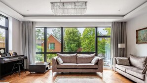 A modern living room with a large window, a sofa, a coffee table, and a desk with a computer. - Highgate - Luxury 4-Bedroom Spacious House with Indoor Pool (London)