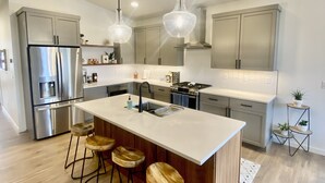 A modern kitchen with a central island, stainless steel appliances, pendant lighting, and wooden stools. - Clean, Comfortable, and Modern CDA Retreat inside Gated Community. (Coeur d'Alene)