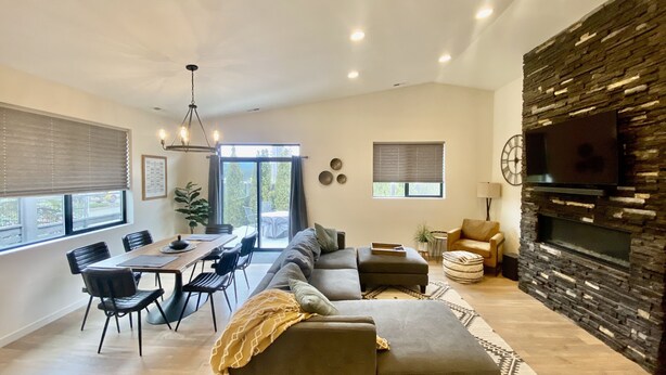 A modern living room with a sectional sofa, a dining area with a table and chairs, a fireplace, and a large TV mounted on a stone wall. - Clean, Comfortable, and Modern CDA Retreat inside Gated Community. (Coeur d'Alene)