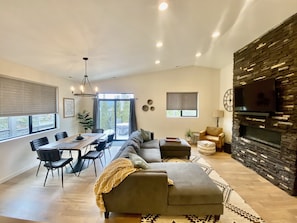 Ein modernes Wohnzimmer mit einer Eckcouch, einem Essbereich mit Tisch und Stühlen, einem Kamin und einem großen Fernseher, der an einer Steinwand befestigt ist.
