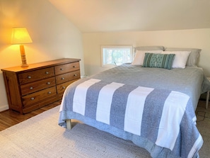 Un dormitorio con cama, cómoda de madera, lámpara y ventana.