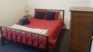 A single bed with a red bedspread, two pillows, a checkered blanket, and a rolled-up towel on the bed. There is a small table with a lamp beside the bed.