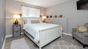 Une chambre à coucher avec un lit blanc, deux tables de chevet avec des lampes, un canapé gris et un téléviseur fixé au mur.