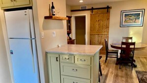 Una cocina con un refrigerador blanco, una isla verde claro con cajones, piso de madera y una zona de comedor con mesa y sillas.