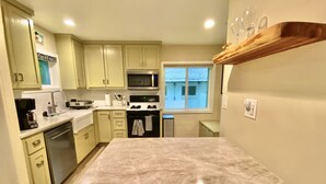 Una cocina con armarios verde claro, un microondas de acero inoxidable, una estufa y un lavavajillas. Hay una ventana que ofrece vistas a una casa.