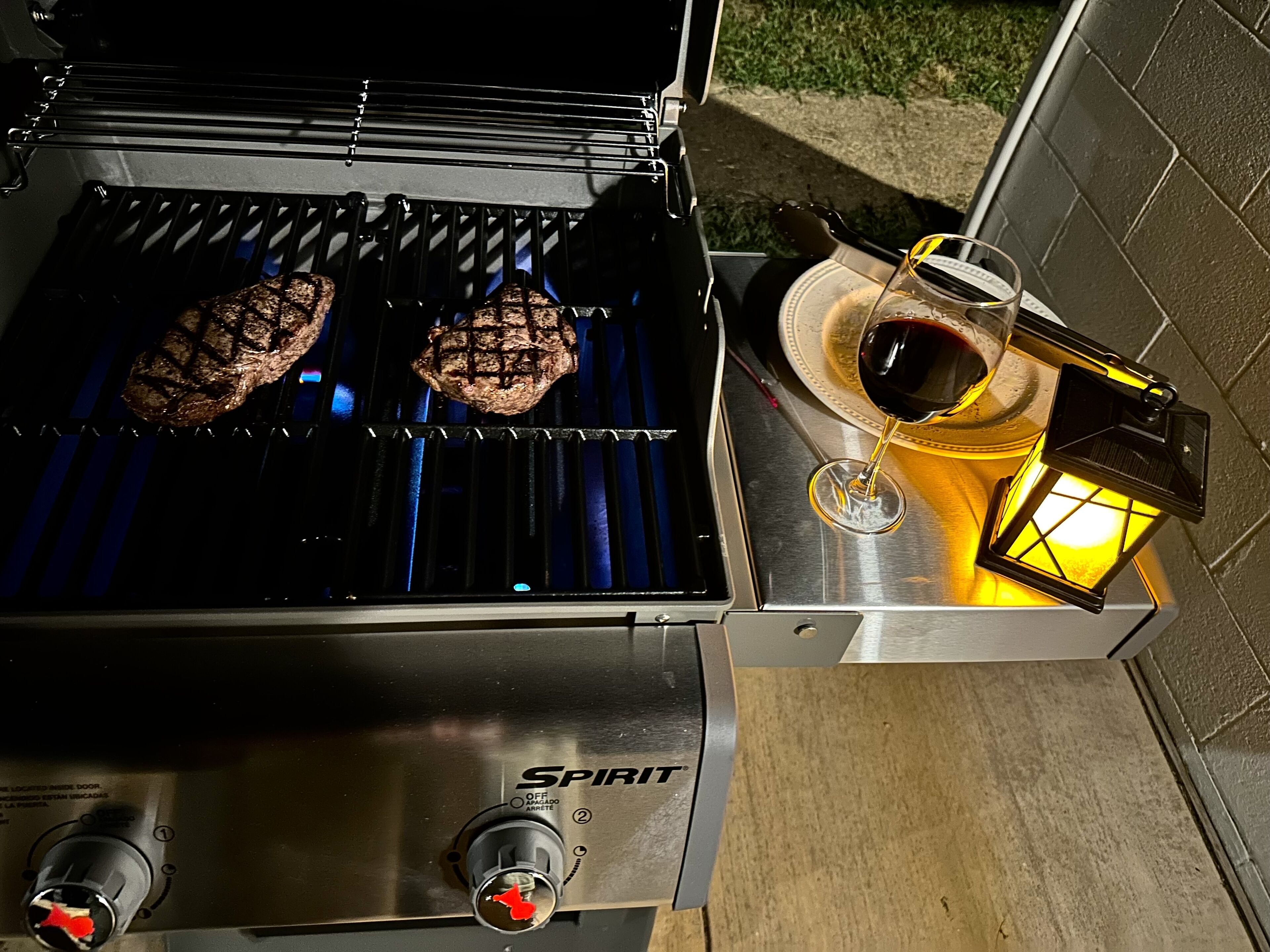 A grill with two steaks cooking, a glass of red wine, and a lantern on a table.