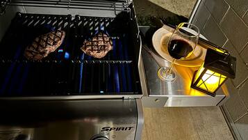 A grill with two steaks cooking, a glass of red wine, and a lantern on a table.