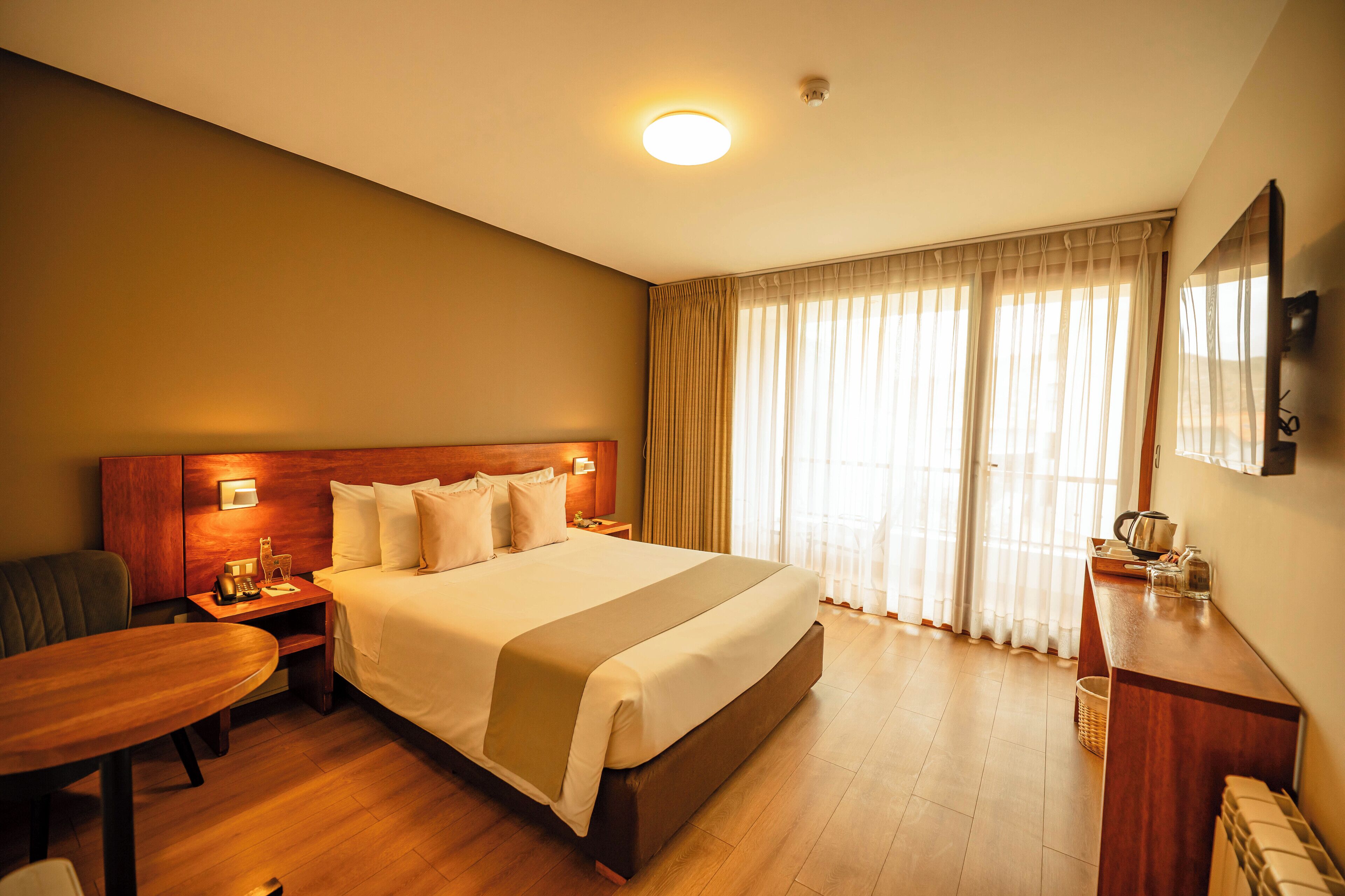 A hotel room with a large bed, a desk, a chair, a small table, a nightstand, and a mirror.