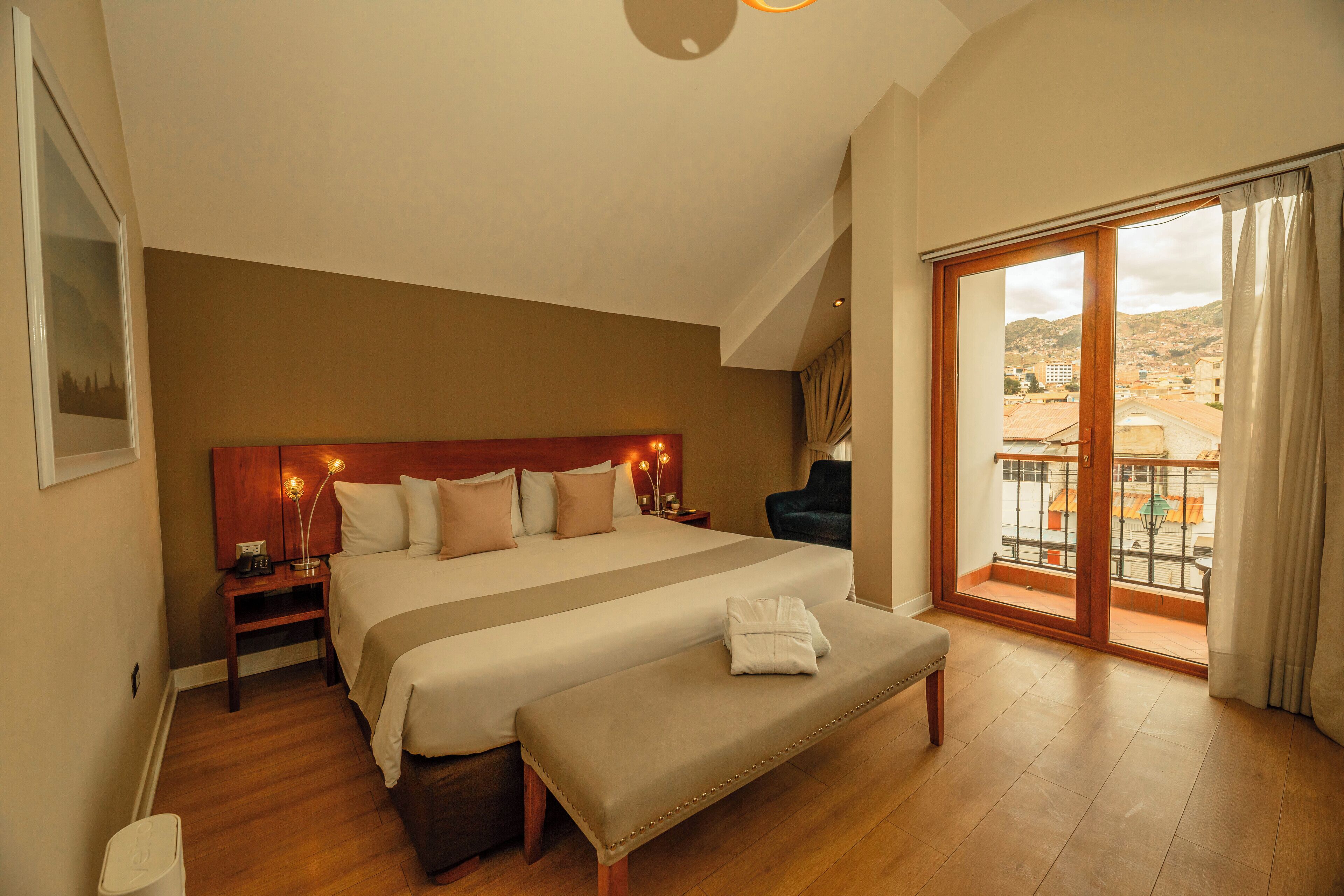 A spacious bedroom with a large bed, a bench, a nightstand, and a view of the city through a balcony.