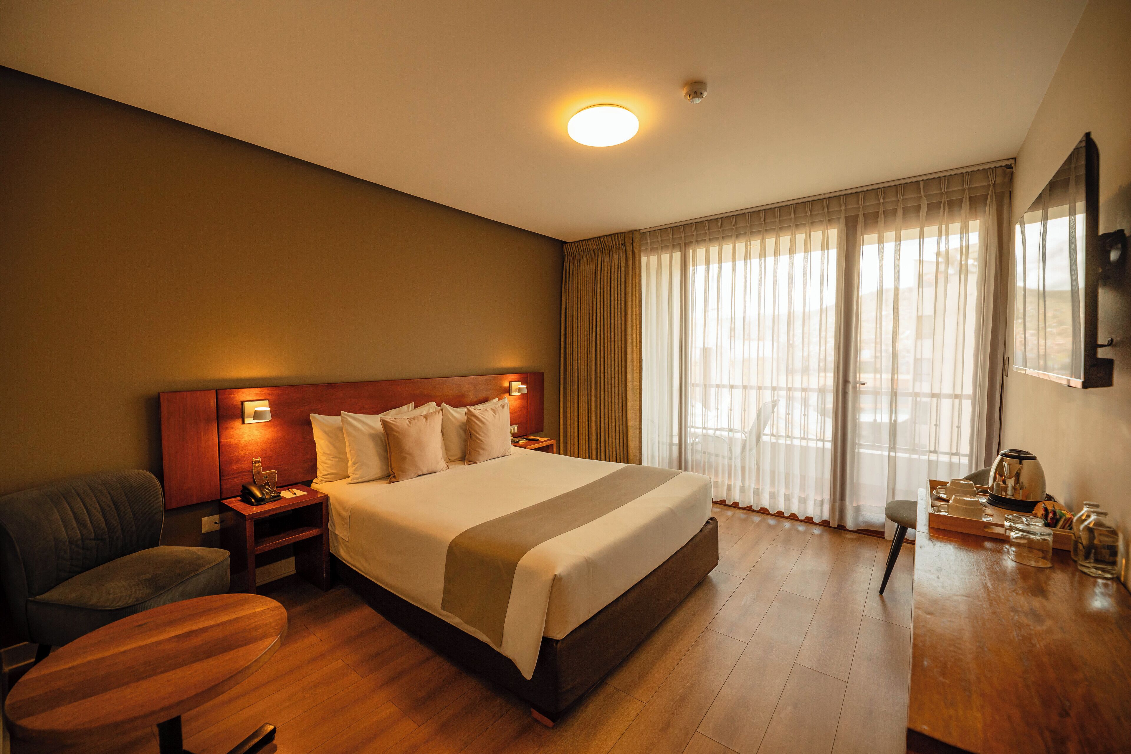 A hotel room with a large bed, a wooden desk, a chair, a TV, and a window with curtains.