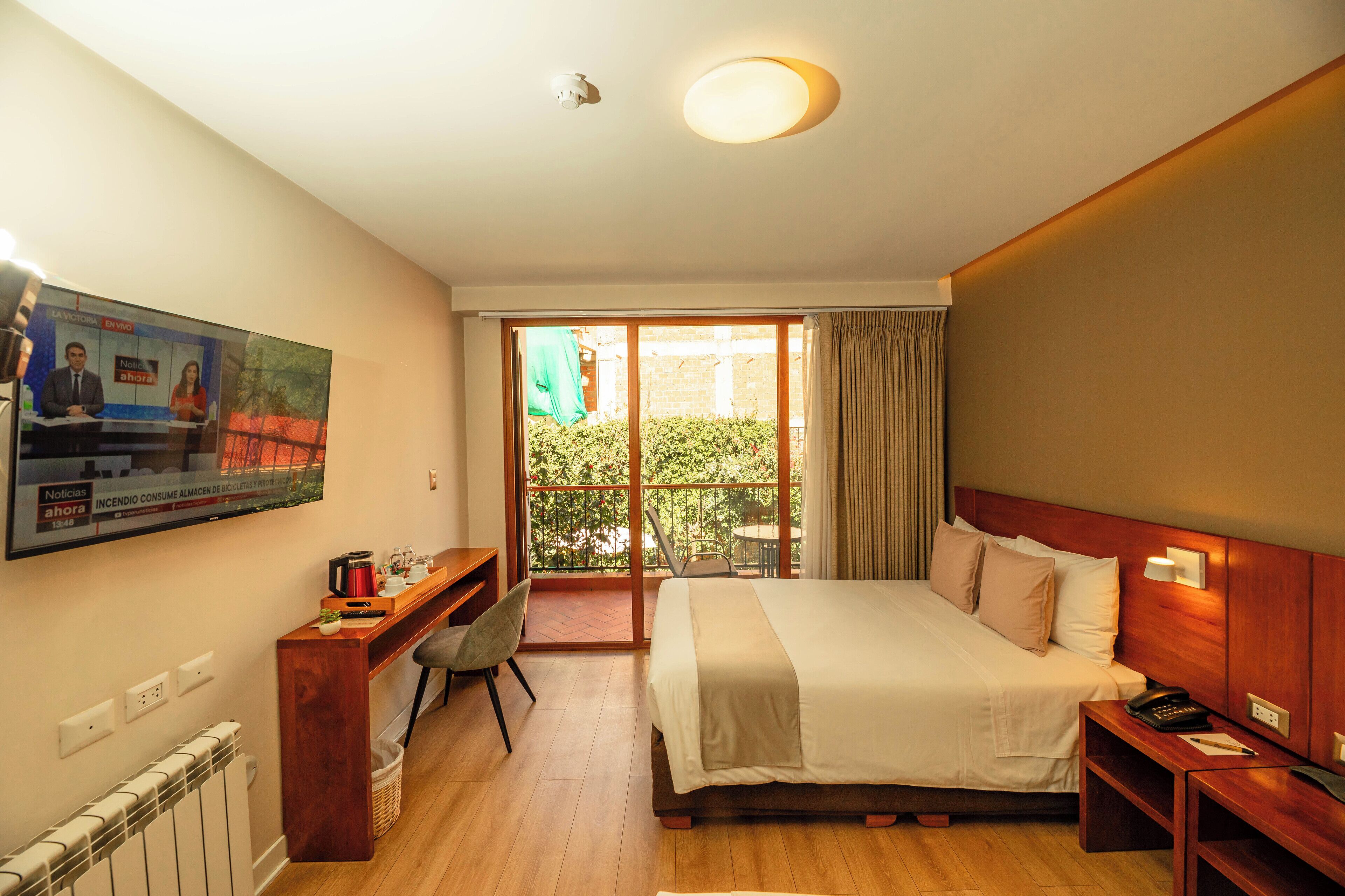 Habitación de hotel con cama, escritorio, televisión y balcón con vista a vegetación.