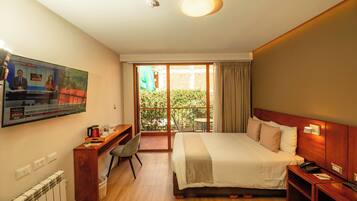Habitación de hotel con cama, escritorio, televisión y balcón con vista a vegetación.