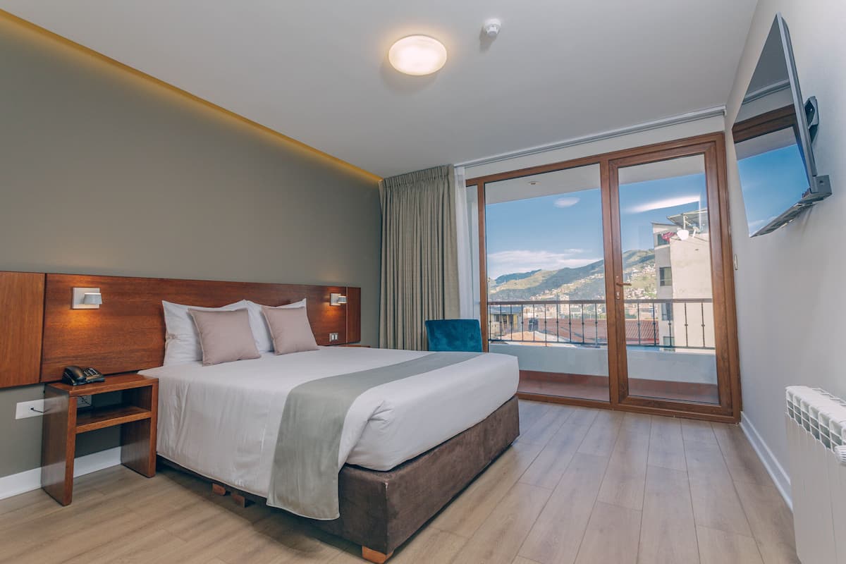 Ein Hotelzimmer mit einem großen Bett, einem Nachttisch mit Telefon, einem an der Wand montierten Fernseher und einem Balkon mit Blick auf die Stadt.