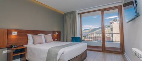 A hotel room with a large bed, a bedside table with a phone, a TV mounted on the wall, and a balcony with a view of the city.