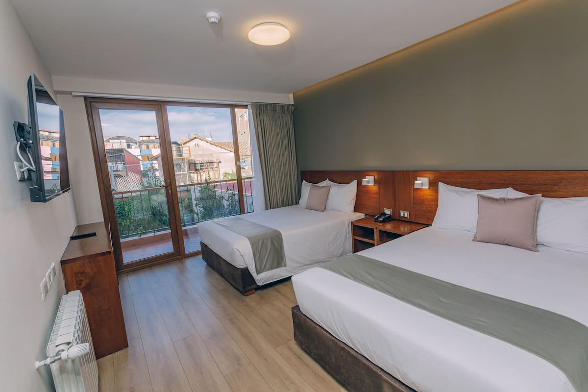 Ein Hotelzimmer mit zwei Betten, einem großen Fenster mit Blick auf Gebäude, einem hölzernen Kopfteil und einem Nachttisch mit Lampe.